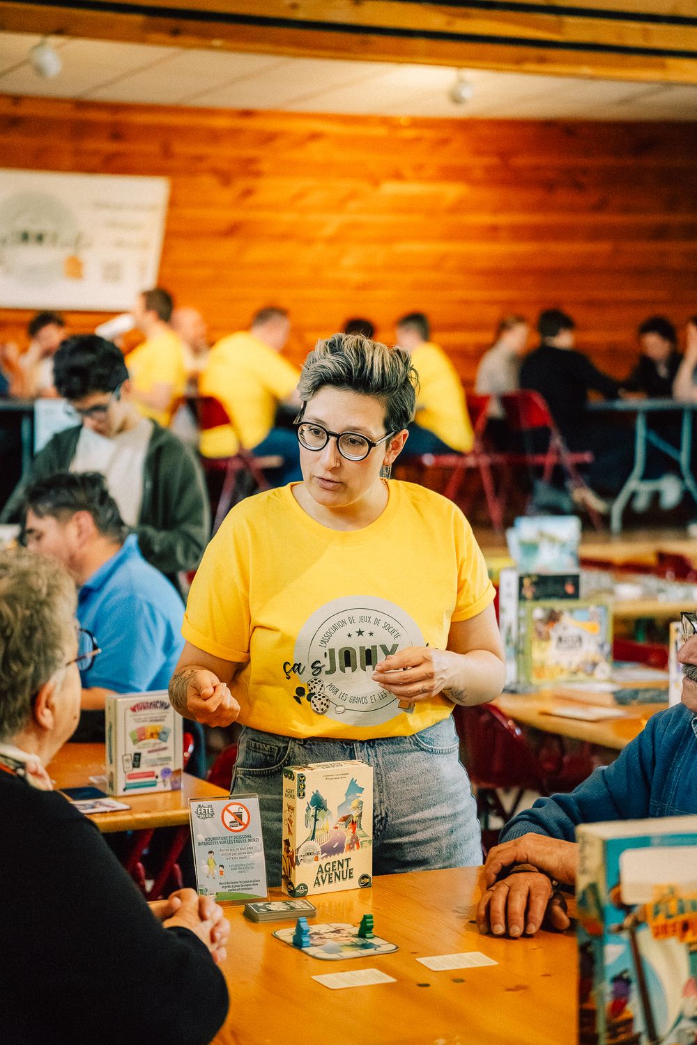 Une membre de l'équipe, en t-shirt jaune Ça s'Joux Vite, explique un jeu à une table